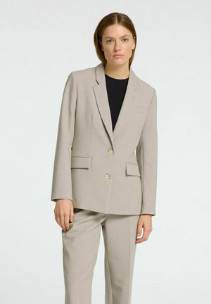 Veste de costume beige avec deux boutons, revers crantés et deux poches avant, assortie à un pantalon. Texture lisse, coupe ajustée.