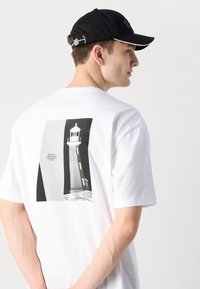 Weißes Baumwoll-T-Shirt mit einem Leuchtturm-Graphic auf der Rückseite. Schwarze Kappe mit dezentem Branding und einem kontrastierenden weißen Saum.