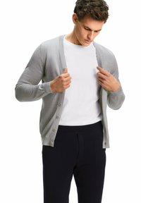 Cardigan grigio chiaro con fronte abbottonato, texture morbida, vestibilità slim e maniche lunghe, indossato sopra una camicia bianca e abbinato a pantaloni scuri.