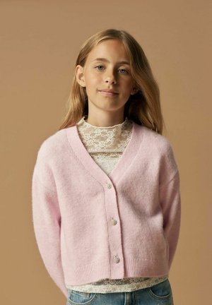Cardigan rose clair et duveteux avec un décolleté en V et trois boutons décoratifs. Porté par-dessus un haut en dentelle blanche. L'arrière-plan est d'un beige neutre.