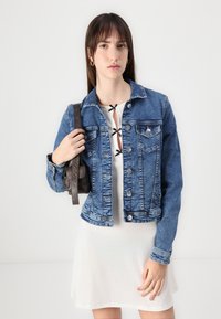 Lys blå denimjakke med metallknapper og to brystlommer. Bært over en hvit kjole med svarte blonder detaljer.