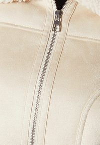 Giacca in pile beige con zip argentata, morbida al tatto e colletto foderato. Cuciture visibili e dettagli del tessuto lisci.