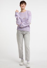 Lila gestrickter Pullover mit strukturiertem Stoff und bunten Pom-Pom-Details. Kombiniert mit hellgrauen Jeans und weißen Sneakers. Einfaches, lässiges Design.
