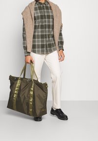 Grüne Nylon-Tragetasche mit olivfarbenen Henkeln, großer rechteckiger Form, glatter Textur und Reißverschluss, gehalten von einer Person, die ein kariertes Hemd trägt.