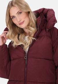 Jachetă puffer bordeaux cu guler înalt, închidere cu fermoar și secțiuni cusute orizontal. Materialul pare neted și căptușit pentru căldură.