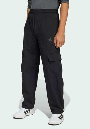 Mann trägt schwarze Cargohosen mit Seitentaschen und weiße Turnschuhe mit schwarzen Streifen, steht vor einem schlichten Hintergrund.