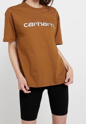 Vrouw draagt een bruin Carhartt T-shirt en zwarte biker shorts, houdt de zoom van het shirt met beide handen vast.