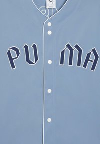 Šviesiai mėlyna beisbolo marškinėlių palaidinė su išsiuvinėtų tekstu "PUMA" tamsiai mėlyna spalva, su baltais akcentais ir penkiomis baltomis spaustukais priekyje.