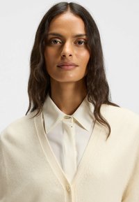 Cream soft cardigan with a V-neck, featuring a textured pattern, worn over a white button-up shirt with a pointed collar.