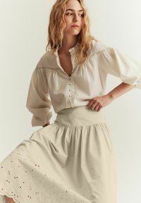 Vit, knapp-up blus med texturerad tyg och puffärmar, i kombination med en beige, lager-på-lager, broderad kjol med blommiga utskärningar.