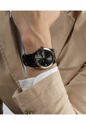 Black watch with a round face, silver accents, and a date display, mounted on a black leather strap, worn on a bare wrist.