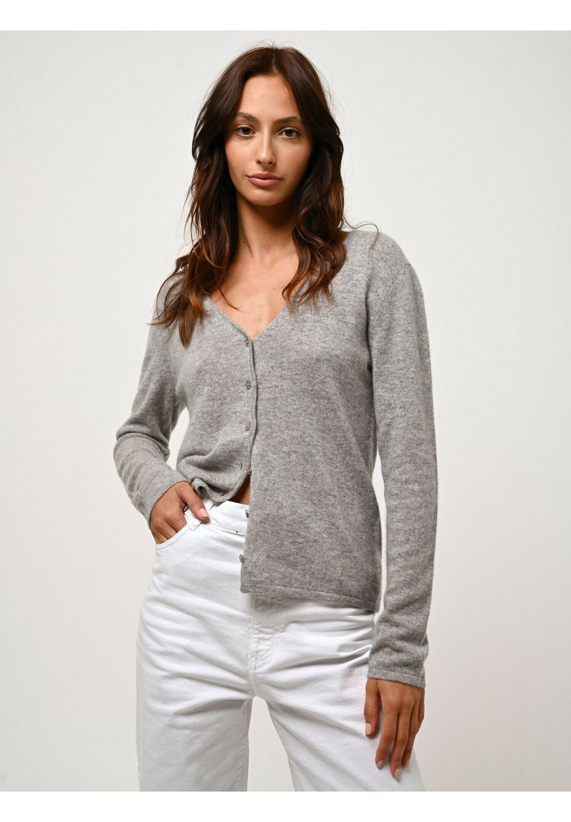 Cardigan grigio realizzato in materiale morbido, con scollatura a V, maniche lunghe e chiusura con bottoni, abbinato a pantaloni bianchi.