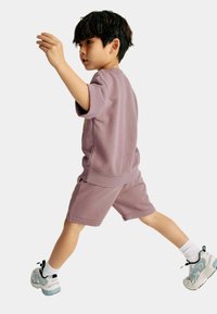 Camiseta de algodón morada con mangas cortas y pantalones cortos a juego. El niño lleva calcetines blancos y zapatillas deportivas con acentos en azul y negro.