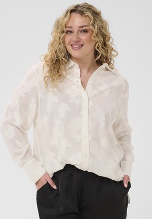 Femme souriante aux cheveux blonds bouclés portant un chemisier blanc transparent à motifs avec boutons et un pantalon noir, les mains dans les poches.