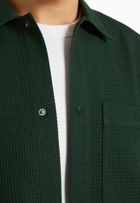 Tmavě zelená texturovaná košile s knoflíkovým zapínáním, našitou kapsou a kontrastním bílým podkošilím. Tkanina má vaflový vzor.