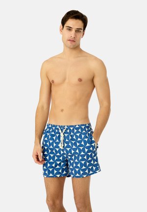 Jeune homme debout, main dans la poche, portant un short de bain à motifs bleus et blancs, sur un fond uni.