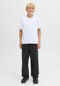 Un garçon blond souriant, portant une chemise blanche à manches courtes unie, un pantalon noir ample et des baskets noires, se tenant devant un fond uni.
