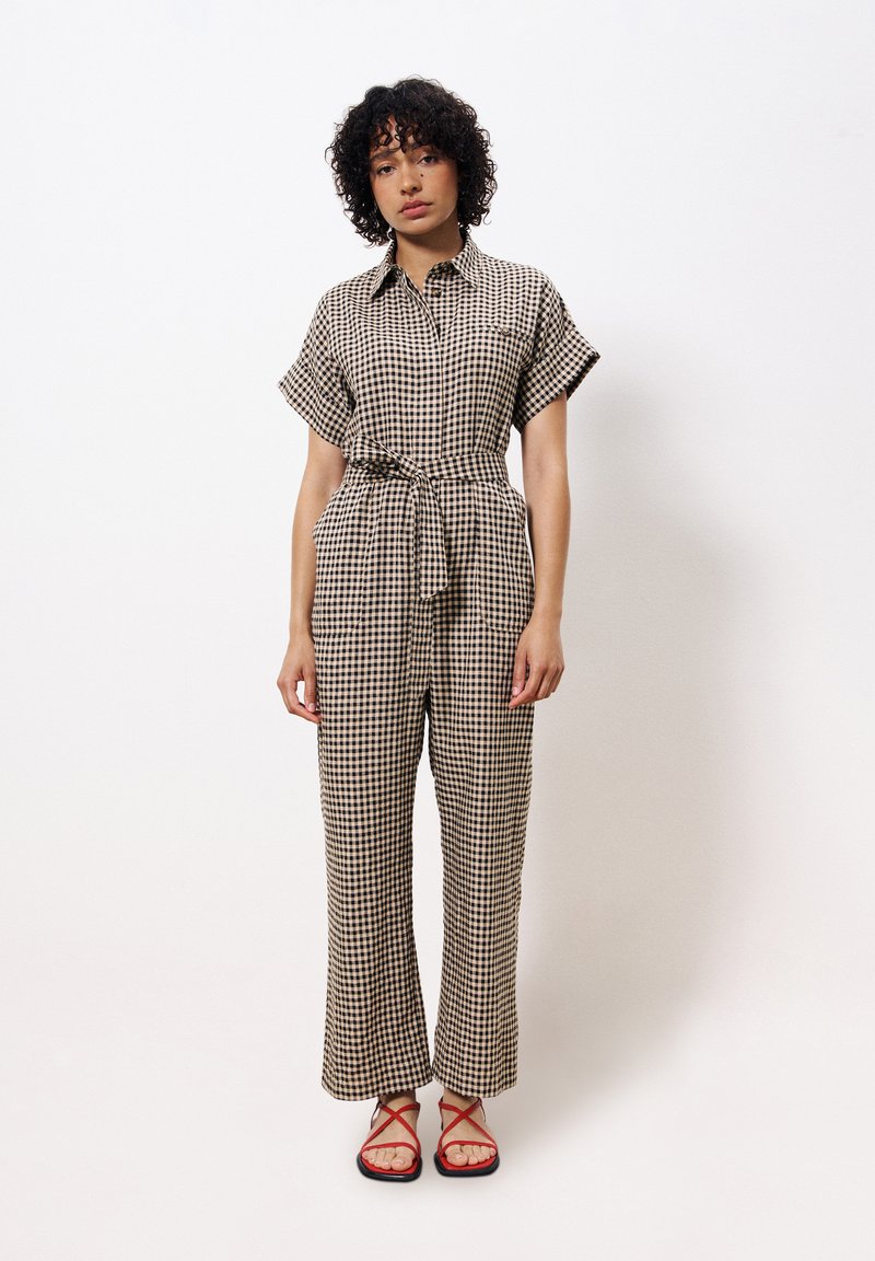 Beige en bruine geruite jumpsuit met korte mouwen, kraag, tailleband en wijde pijpen. Gdragen met rode sandalen met banden.