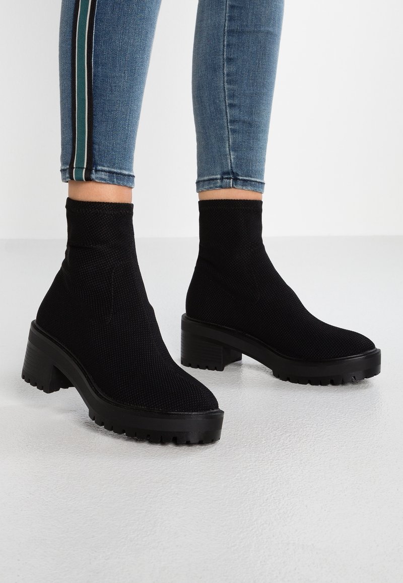 Personne portant des bottines noires texturées à talons épais et semelles crantées, associées à un jean bleu avec des bandes latérales.