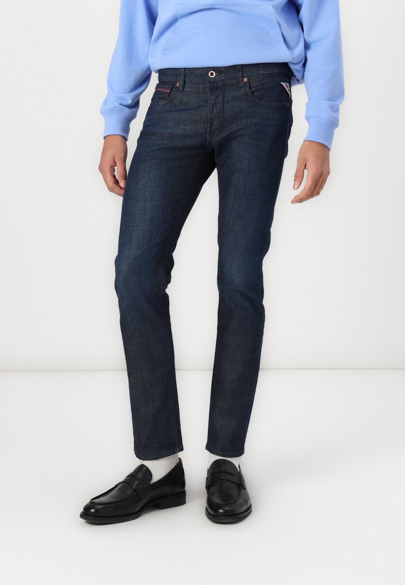 Personne portant un jean bleu foncé, des mocassins noirs avec des chaussettes blanches, et un sweat-shirt bleu clair sur fond blanc.