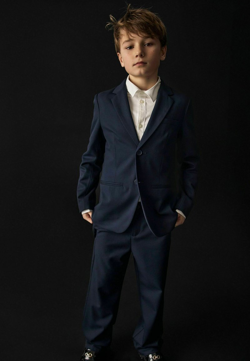 Un jeune garçon se tient debout, les mains dans les poches, portant un costume bleu foncé et une chemise blanche sur un fond noir.