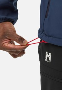 Navy jacket with red adjustment cords at the hem, featuring a hand gripping the cords. Black pants with a white "Millet" logo visible.