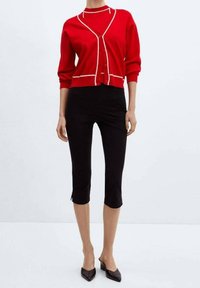 Frau trägt eine rote, geknöpfte Strickjacke mit weißem Rand, schwarze, verkürzte Hosen und schwarze spitze, flache Schuhe, steht vor einem einfarbigen Hintergrund.