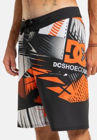Black swim shorts featuring an abstract design in orange, white, and gray, with a drawstring waist and the logo "DCSHOECO." on the front.