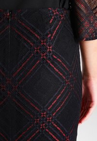 Svart spetskjol med diamantmönster och röda accenter, strukturerad textur och dragkedja bak för stängning.