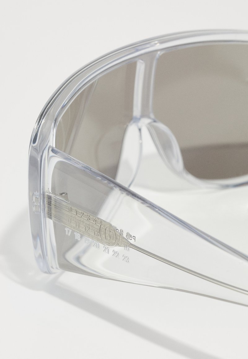 Klare Sonnenbrille mit grauen Gläsern, aus transparentem Rahmen, mit seitlichen Maßen, die auf dem Arm eingraviert sind, und einer glatten, glänzenden Oberfläche.