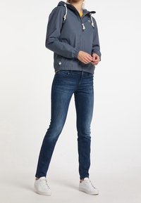 Coupe-vent léger gris avec capuche ajustable, poignets élastiques et deux poches latérales ; porté avec un jean bleu et des baskets blanches.