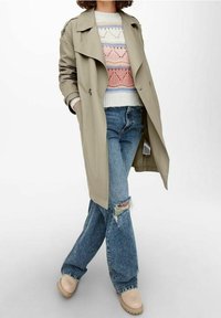 Trench-coat beige, pull en maille avec motifs pastel, jean bleu usé et sabots de couleur claire, présenté en pose debout.