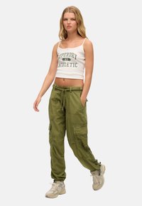 Witte cropped tanktop met groene grafische tekst, gecombineerd met olijfgroene cargobroek met zakken en een trekkoord in de taille, gedragen met beige sneakers.