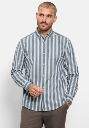 Mann mit kurzen Haaren, der ein blau-weiß vertikal gestreiftes Hemd mit Knöpfen und braune Hosen trägt, schaut seitlich, mit einer Hand in der Hosentasche.