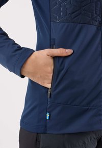 Giacca sportiva blu navy con pannello frontale testurizzato e tasca con cerniera, caratterizzata da un piccolo dettaglio con la bandiera svedese. Realizzata in materiale liscio e elastico.