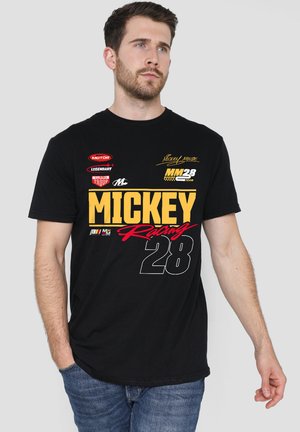 Mann trägt schwarzes T-Shirt mit gelben, roten und weißen Mickey Racing 28 Logos und blaue Jeans, steht mit einer Hand in der Tasche.