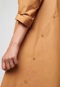 Světle hnědá košile s ohrnutými rukávy, s bočním detailem s knoflíky, hladkou texturou a jednoduchým, elegantním designem.
