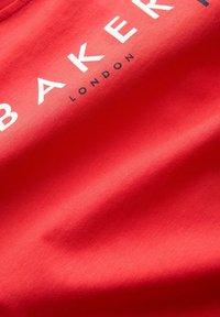 Crvena pamučna majica s plavim tekstom "BAKER" u bijeloj boji, s malim natpisom "LONDON" ispod u tamnoplavoj boji. Glatka tekstura, kružni izrez.
