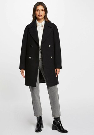 Cappotto nero doppiopetto con ampio colletto a punte, bottoni argentati e silhouette dritta, indossato sopra una camicia bianca e jeans grigi.
