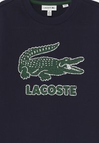 Mörkblå bomullströja med en grön krokodilgrafik med texturerad design och ordet "LACOSTE" under den i matchande grönt.