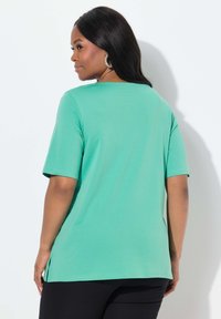 Tyrkysově zelený krátký rukáv top s kulatým výstřihem, se bočními rozparky a hladkým, lehkým materiálem, nošený s černými kalhotami.