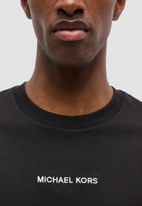 Gros plan d’une personne portant un t-shirt noir avec "MICHAEL KORS" imprimé en blanc sur la poitrine sur un fond uni.