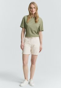 Camiseta verde de manga corta combinada con pantalones cortos color crema que incluyen una cinturilla elástica y puños doblados. Usada con zapatillas blancas.