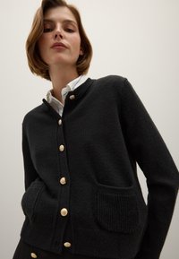 Cardigan noir en tissu tricoté texturé, avec un col rond, une fermeture à boutons dorés, et deux poches avant.