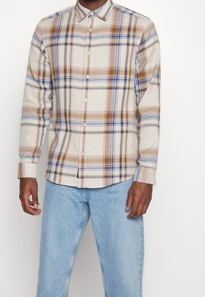 Homme portant une chemise beige à boutons avec un motif à carreaux marron, bleu et noir, associée à un jean bleu clair, sur un fond uni.