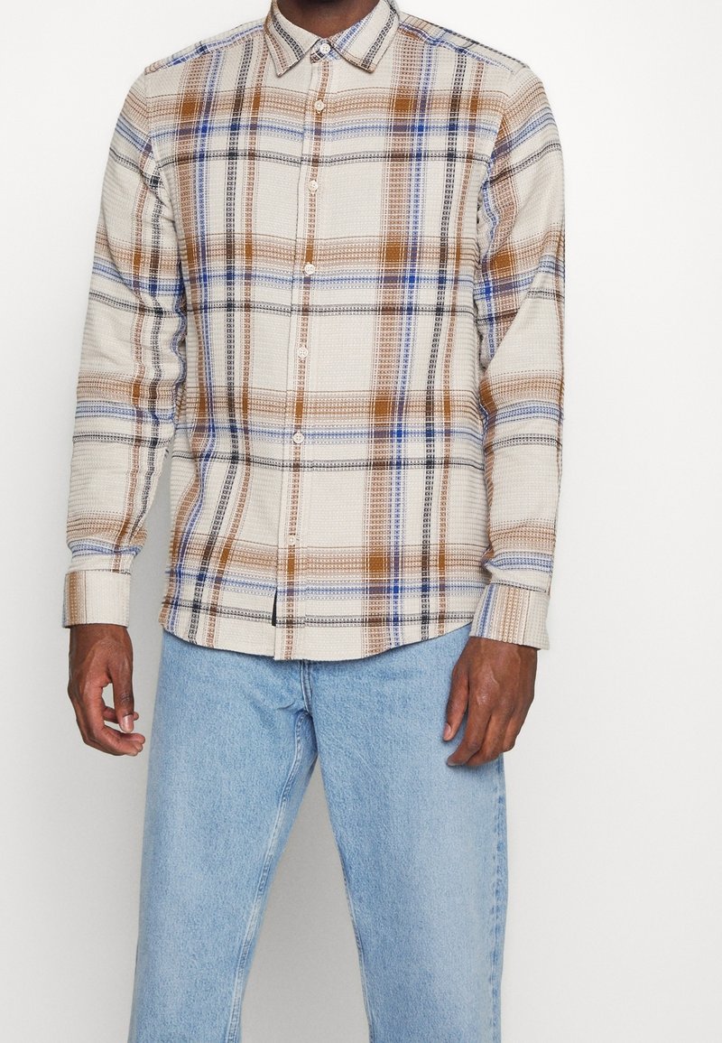 Homme portant une chemise beige à boutons avec un motif à carreaux marron, bleu et noir, associée à un jean bleu clair, sur un fond uni.