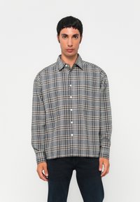 Jonge man in een grijs en wit geruit overhemd met knopen en donkere broek, staande tegen een effen witte achtergrond.