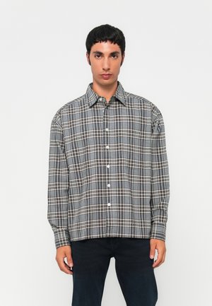 Jonge man in een grijs en wit geruit overhemd met knopen en donkere broek, staande tegen een effen witte achtergrond.