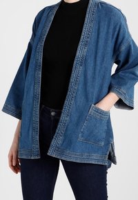 Chaqueta de denim de corte relajado en azul medio. Presenta mangas anchas, frente abierto, bolsillo con parche y costuras visibles. Lleva puesta una camiseta negra.