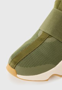 Grüner Mesh-Sportschuh mit einer dicken, strukturierten Gummisohle, der ein breites elastisches Band über dem Obermaterial für einen sicheren Halt aufweist.